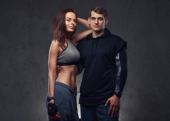 Attractive couple, slim brunette female and handsome guy in sportswear cuddling in a studio on a dark textured background.