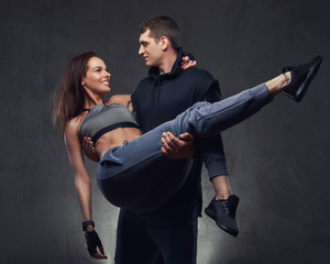 Attractive sports couple. Handsome guy holding his girlfriend on his hands wearing sportswear. Isolated on a dark textured background.