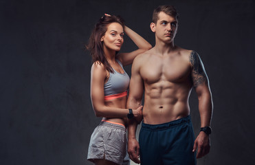 Fototapeta na wymiar Attractive couple, a slim brunette female wearing sports bra and shorts and handsome shirtless guy cuddling in a studio on a dark textured background.