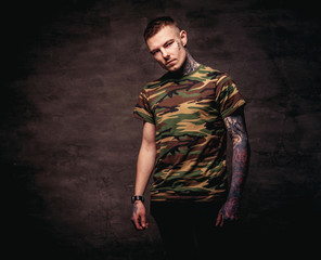 Tattoed young guy in a military t-shirt pose at the studio. Isolated on dark textured background.