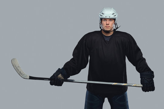 Hockey Player Wearing Black Protective Gear And White Helmet Holds A Hockey Stick On A Gray Background.