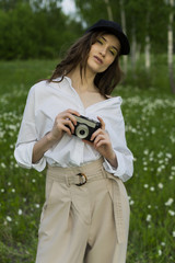 Beautiful girl photographer wearing a white shirt, beige trousers poses outdoors with a vintage camera against a background of meadow full of wild flowers