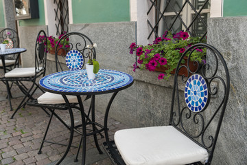 Chairs and tables in a street, decorated with flowers. House wall with flowers in the background.