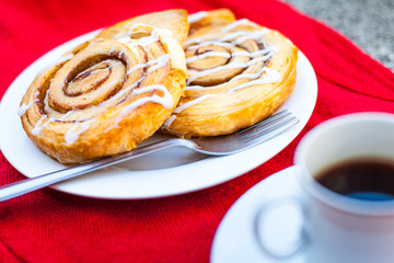 Two cinnamon pastries on a white plate with a coffee in a white cup and saucer, all on a red cloth