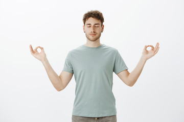 Portrait of calm good-looking mature Caucasian boyfriend in t-shirt, smiling and feeling relaxed, standing with spread hands in zen gesture and closed eyes, meditating or practicing yoga