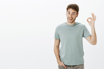 Studio shot of funny friendly-looking european male coworker in trendy t-shirt, raising hand in okay or fine gesture and smiling broadly, giving approval or agreeing with friend opinion