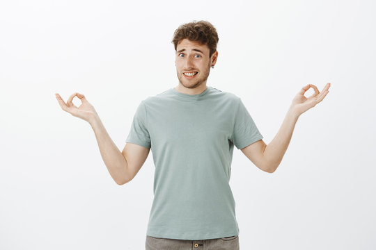 Indoor shot of awkward unsure funny guy with bristle in earrings, smiling clumsy while standing with spread hands in zen gesture and being clueless how meditate or do yoga, standing over gray wall
