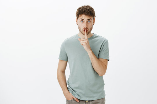 Shut up, do not speak. Portrait of displeased angry male teacher in casual t-shirt, lifting eyebrows from anger, saying shh while showing shush gesture and staring scary at camera, asking keep secret.