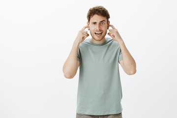 Naklejka premium Indoor shot of funny caucasian boyfriend in casual t-shirt, making faces, covering hearing with index fingers in ears, wanting not listen how girlfriend shouts, posing over gray background