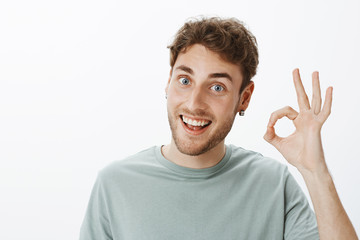 Fine, I got it. Portrait of pleased charming european male with fair hair in earrings, showing ok...