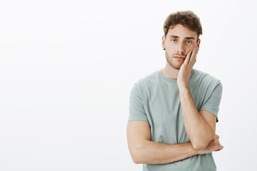 How boring, leave me alone. Portrait of careless handsome guy in earrings, holding hand on palms and staring with indifferent expression at camera, feeling boredom and lack of interest over grey wall