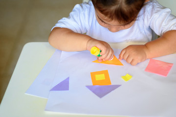 Cute little girl make applique, glues colorful house, applying a color paper using glue stick while doing arts and crafts in preschool or home. The idea for children's creativity, art project.