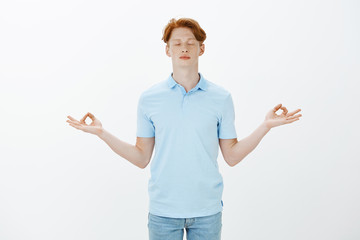 Calm down, need some relaxation. Studio shot of handsome redhead male student with freckles and tattos, standing with closed eyes and spread hands in zen gesture, meditating or practicing yoga