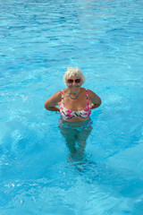 Fototapeta na wymiar Aged woman is standing in bright blue pool water.