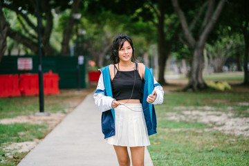 An image of a young Chinese Asian skater girl dancing to the music from her headphones while walking through the park. She is cute, cool, and sporty.