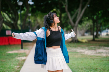 A pretty and young Chinese Asian skater girl is dancing in the park while listening to her music on her headphones. She is wearing casual and trendy clothes.