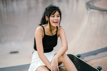 A young Chinese Asian millennial sits at the skate park on a lovely day. She is resting in between skate boarding and listening to music on her wireless headset.