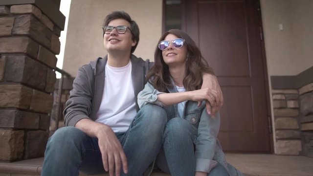 Smiling young hipster guy in glasses is hugging his happy young wife sitting on their house porch. Family values concept. Slider slow motion medium shot