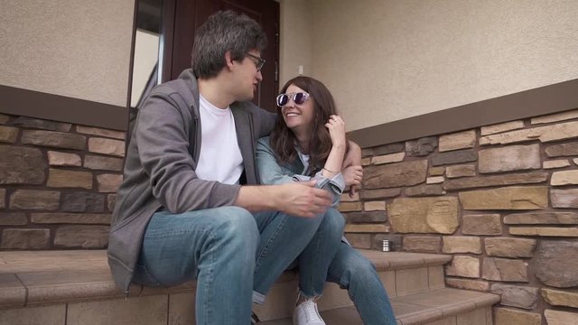 Smiling young hipster guy in glasses is hugging his happy young wife sitting on their house porch. Family values and happy times concept. Slider slow motion medium shot