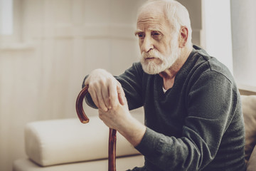 Walking difficulties. Aged bearded man leaning on his walking stick while thinking about his...