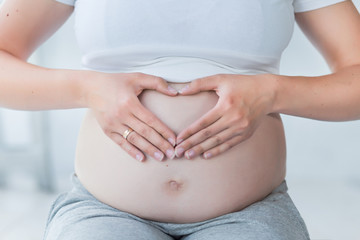 Close-up of pregnant woman holding hands in a heart shape on her belly. Maternity concept. Healthy pregnancy and lifestyle