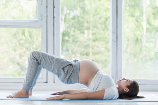 Beautiful young pregnant woman doing yoga or fitness exercise, lotus pose on the blue yoga mat at home. Happy and healthy pregnancy, meditation, calmness concept