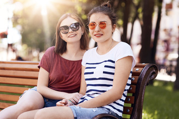 Joyful female lesbians keep hands together, wear trendy sunglasses, enjoy togetherness, sit on...