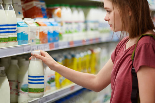 Horizontal view of serious beautiful young female chooses milk products in dairy deparment of supermarket, dressed in casual t shirt, has short hair. People, lifestyle and consumerism concept
