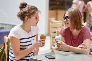 Obraz na płótnie Canvas Indoor shot of happy young female friends look joyfully at each other, eat ice cream, discuss something pleasant with cheerful expressions, sit against cozy cafe interior. Companionship concept