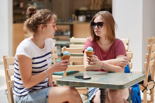 Friends, recreation and summer concept. Two beautiful female teenagers eat cold delicious ice cream, talk with each other, discuss paln for going to sea, enjoy pleasant friendly conversation