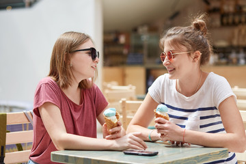 Horizontal shot of friendly females meet together in cafe, eat ice cream and have positive expressions, enjoy summer rest, wears casual t shirts, pose against coffee shop interior. Friendship concept