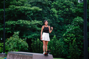 A portrait of a smiling, young, attractive Chinese Asian skater girl walking gracefully in the park. She is ready to use her skateboard and go into the ramp.