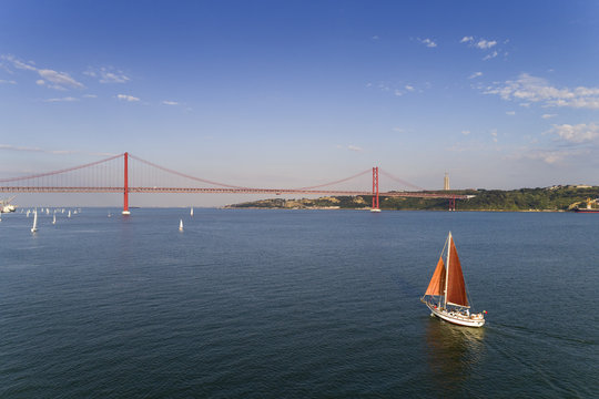 Aerial view of a beautiful sail boat on the Tagus River with the 25 of April Bridge on the background, in the city of Lisbon, Portugal