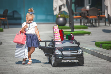 Pretty little girl fashionista making shopping with shopping bags. Cute child outdoors in the toy car