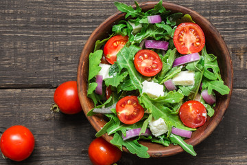 Fresh salad with rucola, tomatoes cherry, feta cheese and red onion in a bowl on rustic wooden table. top view