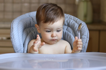 Cute little hungry baby boy holds spoon in his hand and looks at empty table waiting for food, baby gestures