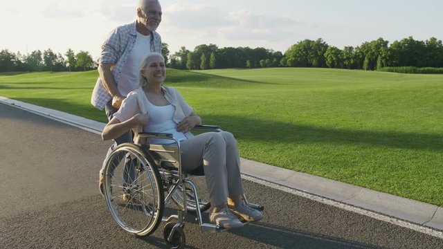 Beautiful senior couple with wife in wheelchair enjoying a walk in summer park. Positive cheerful disabled female kissing and hugging her husband during stroll. Steadicam shot