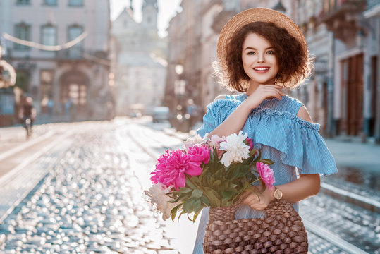 Outdoor portrait of young beautiful happy smiling woman wearing hat, striped dress, holding straw bag with peonies, posing in street of european city. Female fashion concept. Copy space for text 