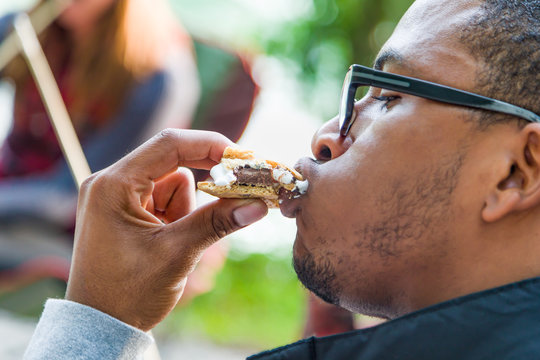 Young Adult African American Man And Friends Enjoys Eating Smore With Roasted Marshmallow And Chocolate At Campfire Outdoors