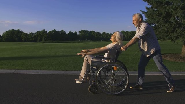Senior handsome bearded man pushing wife in wheelchair while taking a walk in countryside. Positive beautiful senior couple with white hair enjoying and having fun together outdoors. Steadicam