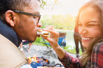 Young Adult Hispanic Girl Feeding African American Man A Smore at Campfire Outdoors