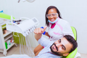 The patient in the dental chair joyfully thumbs up. Snow-white smile. Black African dentist. A modernly equipped medical office.
