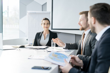 Group of young entrepreneurs sitting at meeting table in conference room and discussing startup projects, focus on smiling woman looking at colleagues, copy space