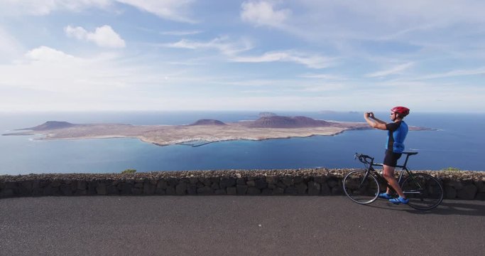 Cyclist tourist taking photo picture with smartphone on biking cycling trip on summer vacation travel. Man riding road bike using phone taking photos on holidays on Lanzarote, Canary Islands, Spain.