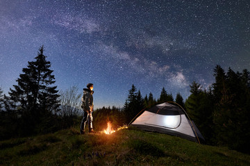 Camping site in mountain valley at night. Young man standing near tourist tent at bonfire under night blue starry sky with Milky way. Beauty of nature, outdoor activity, tourism and traveling concept © anatoliy_gleb