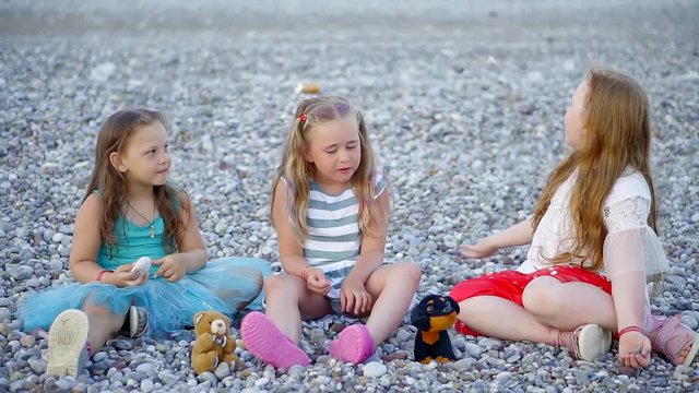 three cute little girl are sitting on a gravel beach in evening, playing and chatting to each other