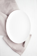 Empty white oval plate on wooden table with linen napkin. Overhead view.
