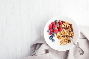 Granola with yogurt and berries in bowl on the white wooden table decorated with napkin. Top view.
