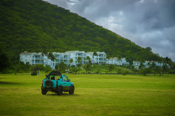 Fototapeta premium Green jeep on green grass field with mountain and white houses in background with blue sky