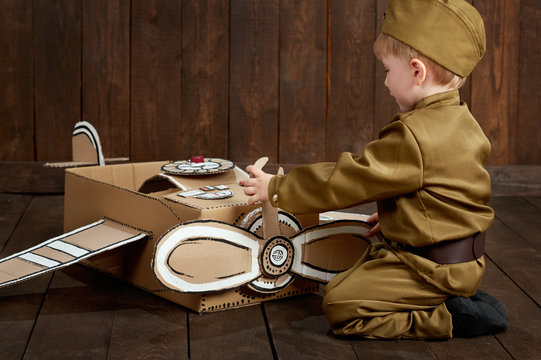 Children Boy Are Dressed As Soldier In Retro Military Uniforms Repair An Airplane Made Of Cardboard Box, Dark Wood Background, Retro Style